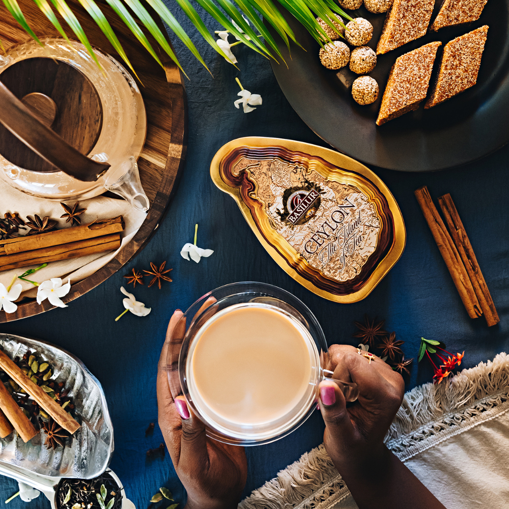 Puszka herbaty Basilur Ceylon otoczona przyprawami, słodkościami i filiżanką mlecznej herbaty trzymaną w dłoniach.