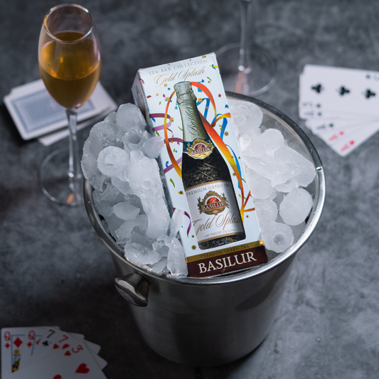 Opakowanie herbaty Basilur Gold Splash w wiaderku z lodem, obok kieliszek napoju i karty do gry na eleganckim stole.
