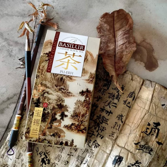 Opakowanie herbaty Basilur Pu Erh na tle chińskiego pergaminu z kaligrafią, pędzlami i suszonym liściem herbaty.