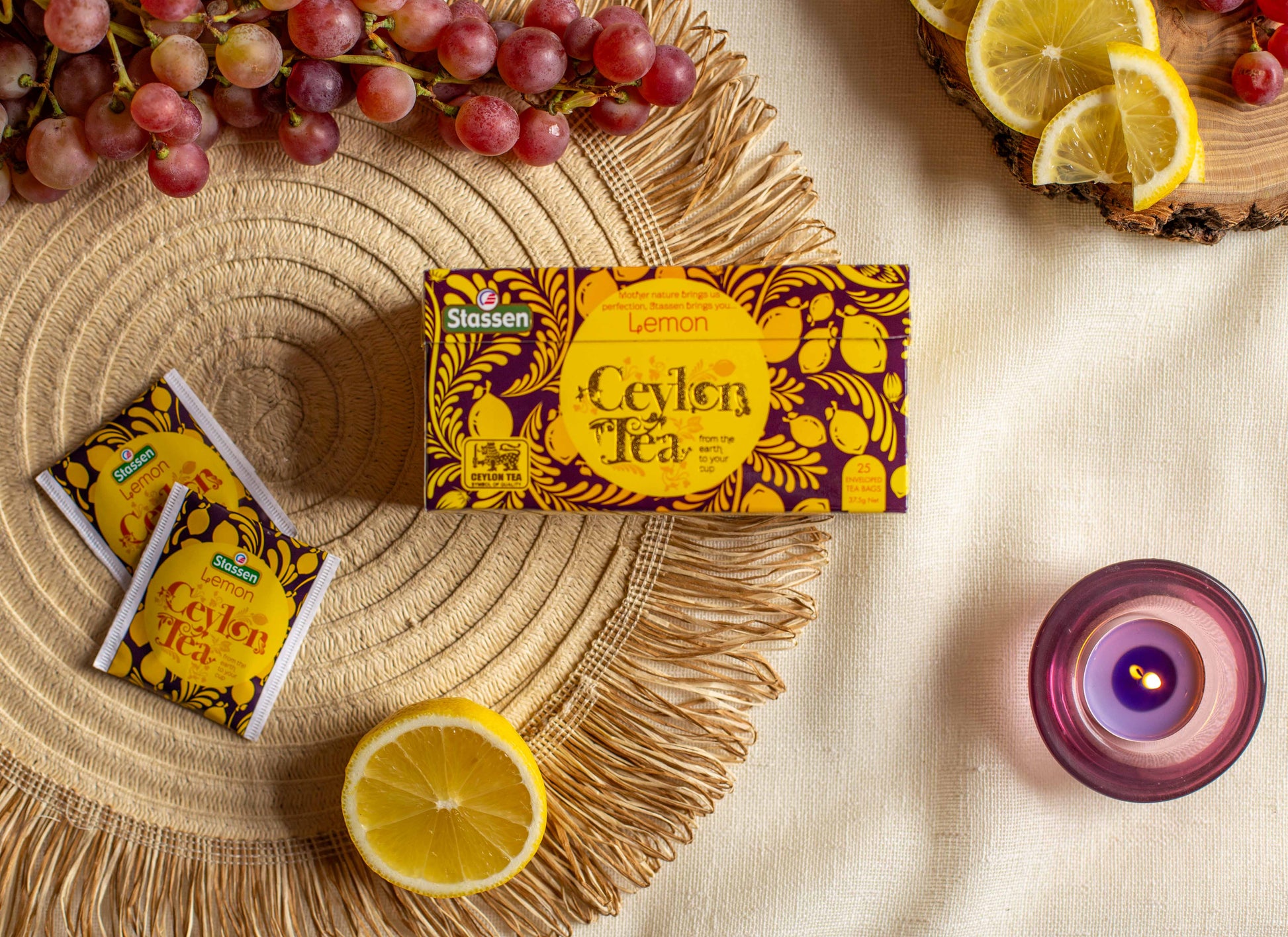 Kompozycja produktowa prezentująca herbatę Stassen Lemon Ceylon Tea na tle plecionki i jasnego obrusu. W centrum znajduje się żółto fioletowe pudełko herbaty o cytrynowym aromacie. Po lewej leżą dwie saszetki w identycznej szacie graficznej oraz połówka cytryny, nad nimi winogrona. Po prawej stronie widoczne są plastry cytryny na drewnianej desce i fioletowa zapalona świeczka. Zdjęcie podkreśla świeżość cytrynowej herbaty Ceylon i ma charakter lifestyle sprzyjający promocji produktu.