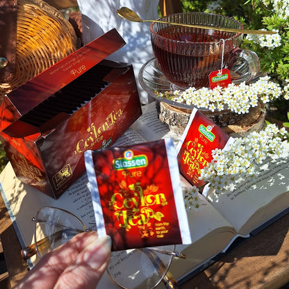 Herbata Stassen Pure Ceylon Tea w saszetkach, szklana filiżanka naparu i otwarta książka wśród białych kwiatów w ogrodzie.