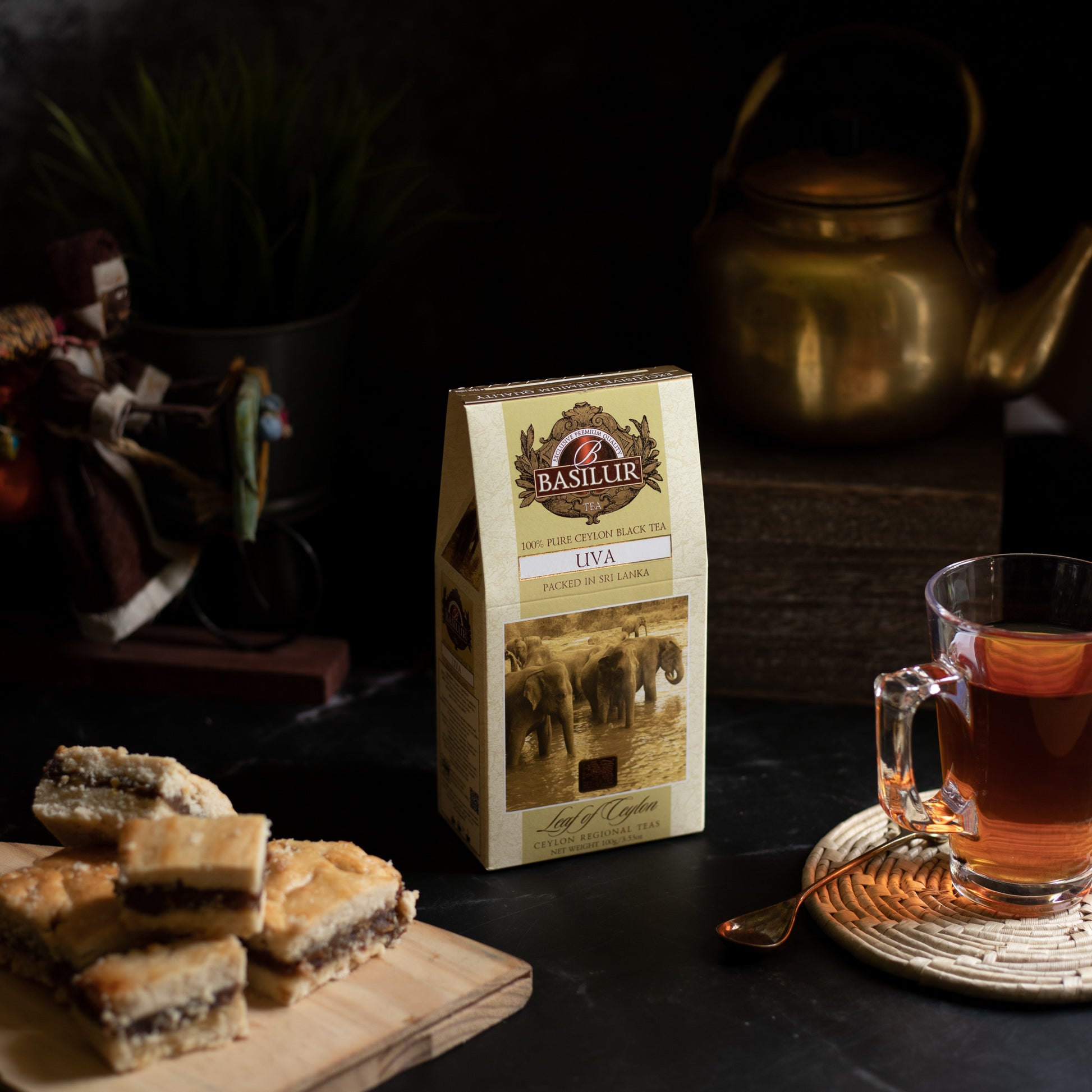 Opakowanie herbaty Basilur Uva ustawione na stole obok szklanki z gorącą herbatą i kawałków ciasta.