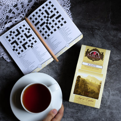 Opakowanie herbaty Basilur Kandy obok filiżanki czarnej herbaty i otwartej książki z krzyżówkami.