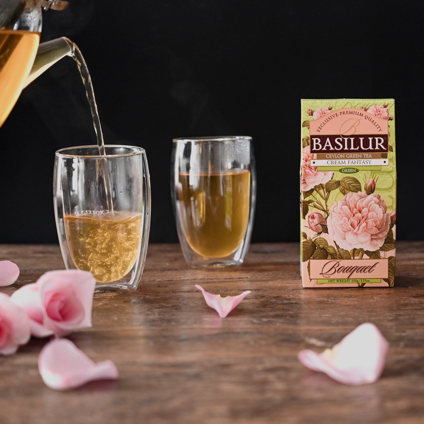 Zielona herbata Basilur Cream Fantasy na drewnianym stole obok dwóch szklanek z gorącym naparem i różanych płatków.