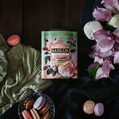 Puszka herbaty Basilur Cream Fantasy otoczona makaronikami i kwiatami na ciemnym tle.