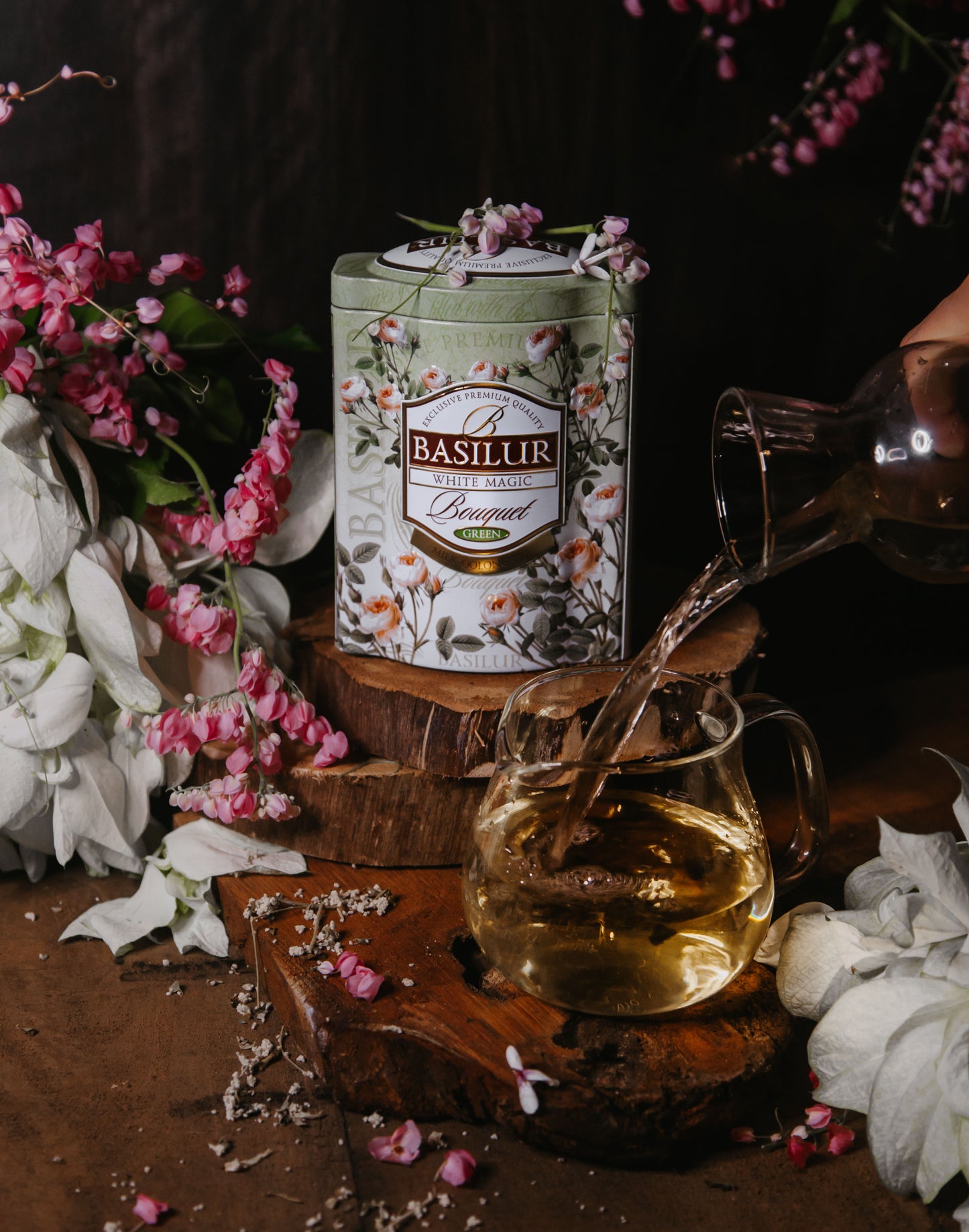 Puszka herbaty Basilur White Magic wśród kwiatów obok szklanki, do której wlewany jest jasny napar.