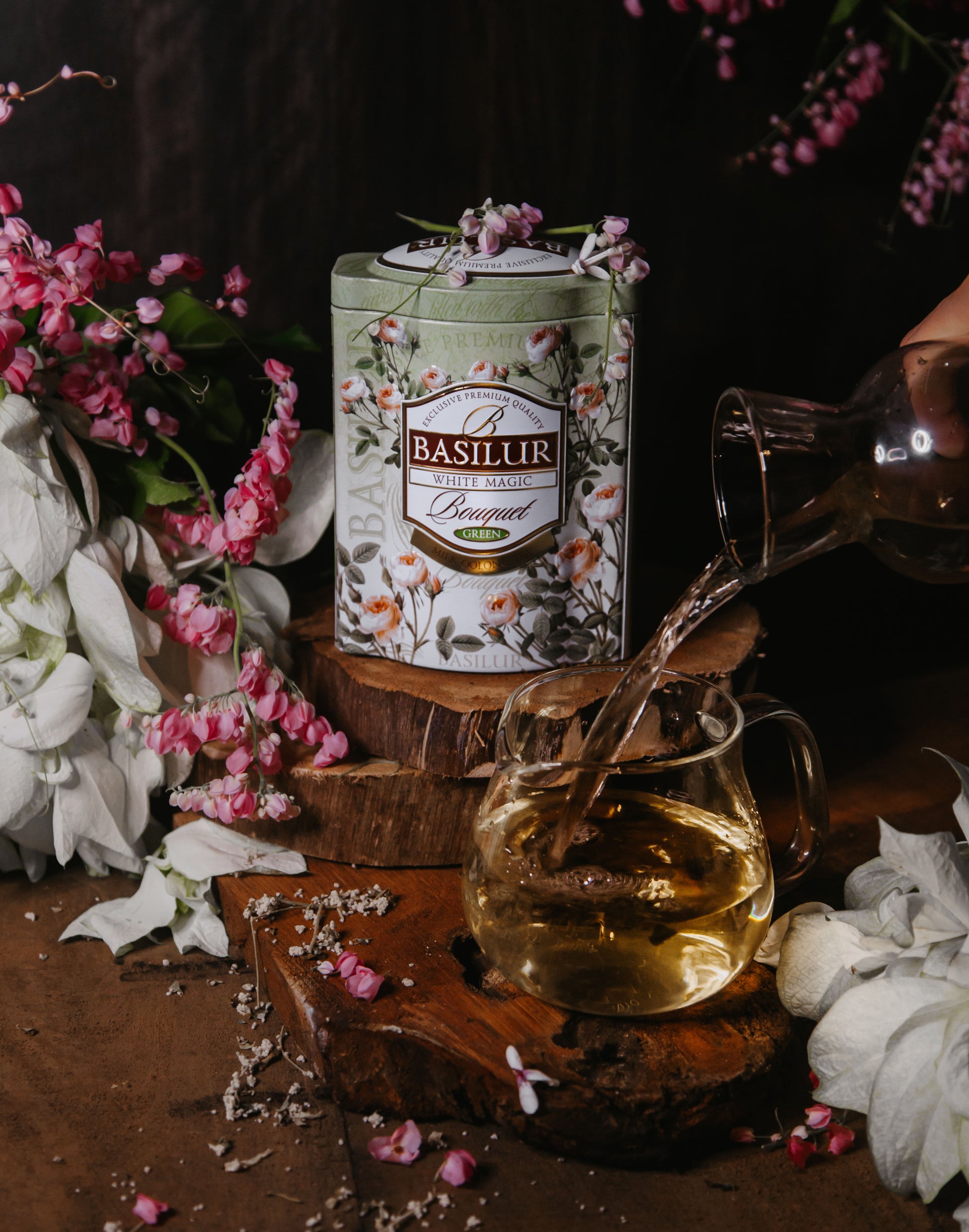 Puszka herbaty Basilur White Magic wśród kwiatów obok szklanki, do której wlewany jest jasny napar.