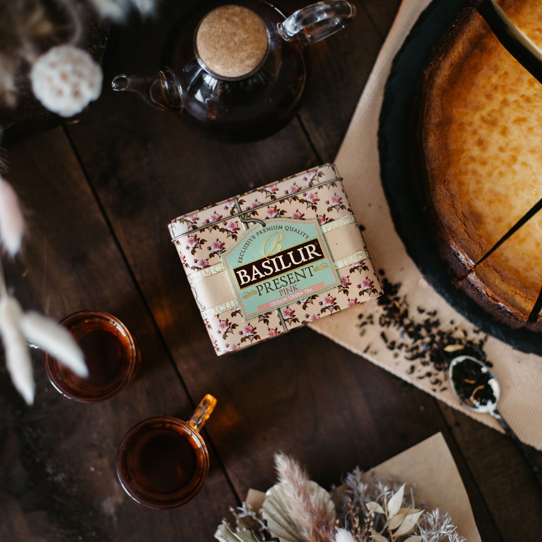 Puszka herbaty Basilur Present Pink na drewnianym stole obok czajniczka, filiżanek i ciasta.