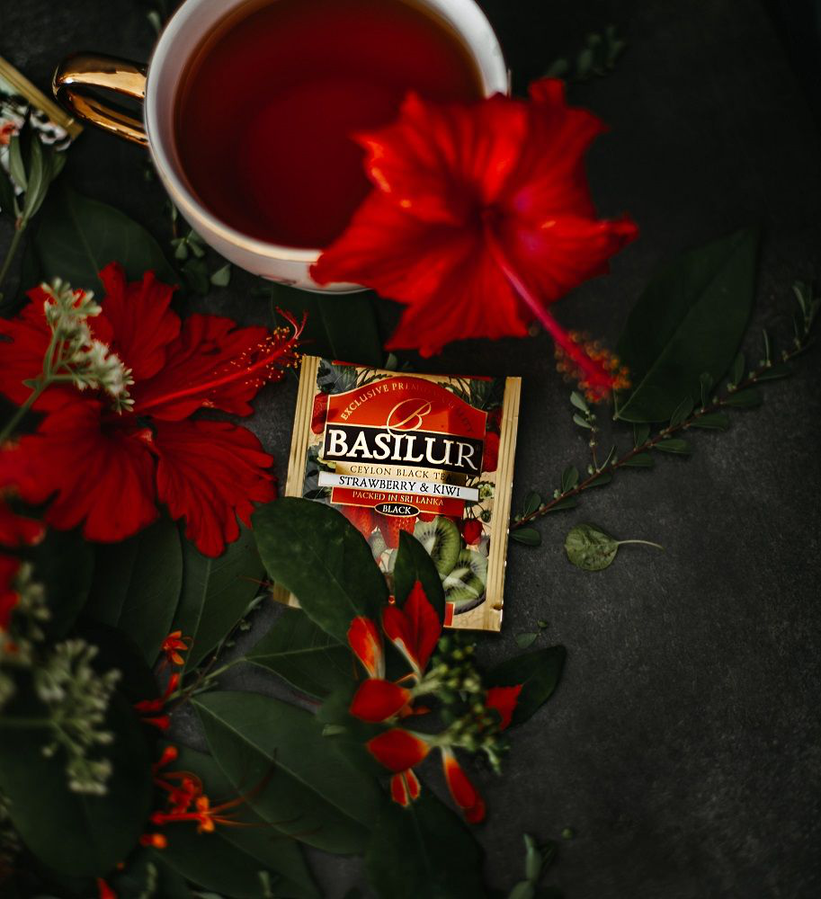 Filiżanka herbaty Basilur Strawberry & Kiwi z czerwoną saszetką i kwiatem hibiskusa.
