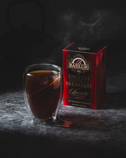 Czerwona herbata Basilur English Breakfast obok szklanki z gorącym naparem na ciemnym tle.