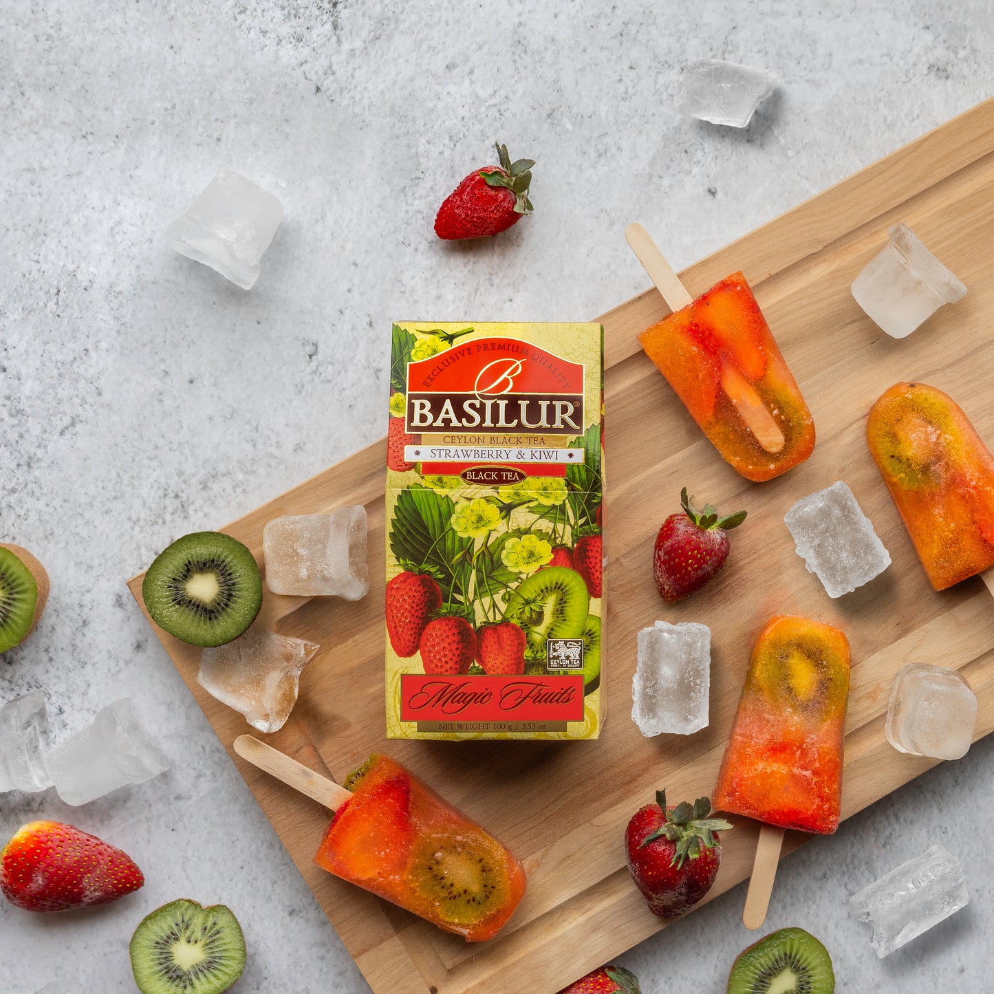 Herbata Basilur Strawberry and Kiwi na desce obok lodów z owocami, truskawek, kiwi i kostek lodu.