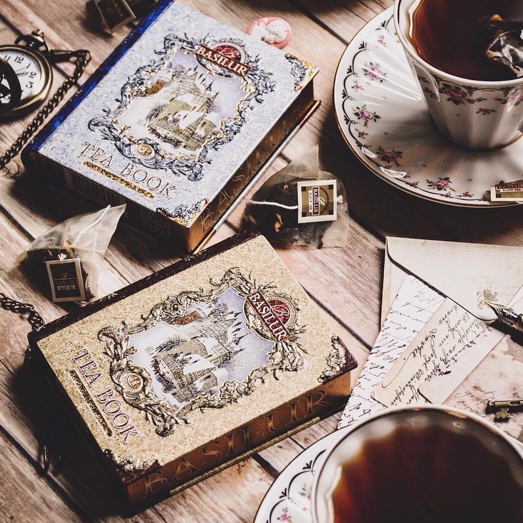 Puszka Basilur Tea Book Vol I w formie książki na drewnianym stole obok filiżanki herbaty i koperty.