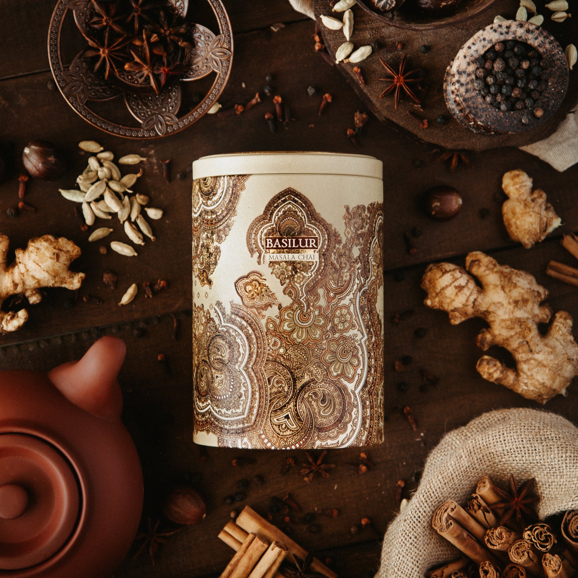 Puszka herbaty Basilur Masala Chai otoczona korzennymi przyprawami jak imbir, kardamon i anyż na drewnianym tle.