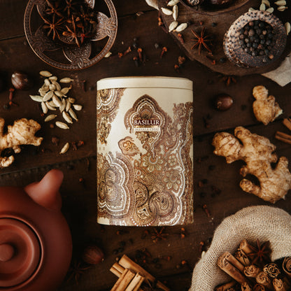 Puszka herbaty Basilur Masala Chai otoczona korzennymi przyprawami jak imbir, kardamon i anyż na drewnianym tle.