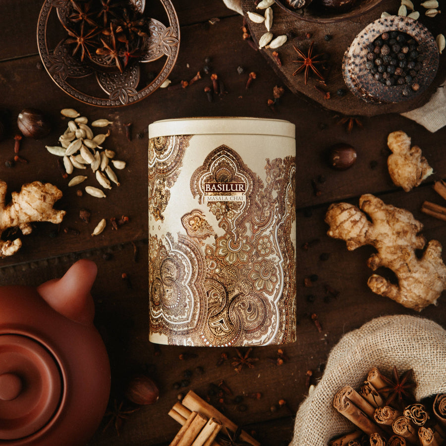 Puszka herbaty Basilur Masala Chai otoczona korzennymi przyprawami jak imbir, kardamon i anyż na drewnianym tle.