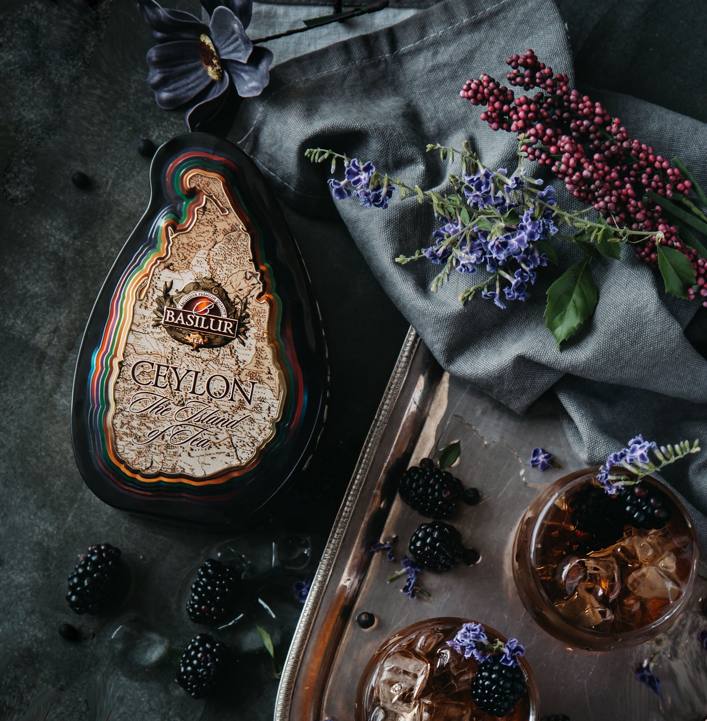 Puszka herbaty Basilur Special w kształcie wyspy Cejlon obok jeżyn i kwiatów na ciemnym tle.