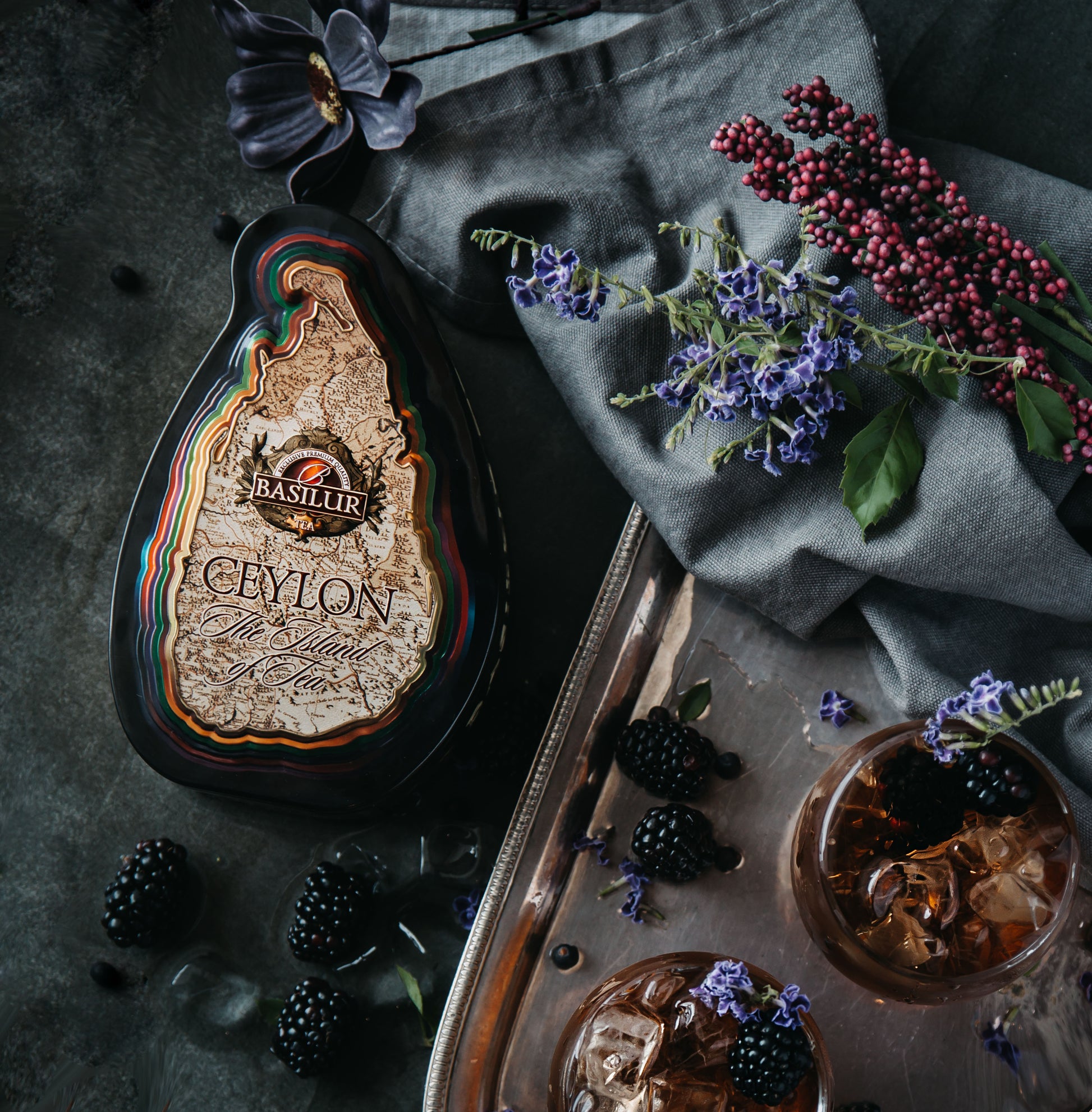 Puszka herbaty Basilur Special w kształcie wyspy Cejlon obok jeżyn i kwiatów na ciemnym tle.