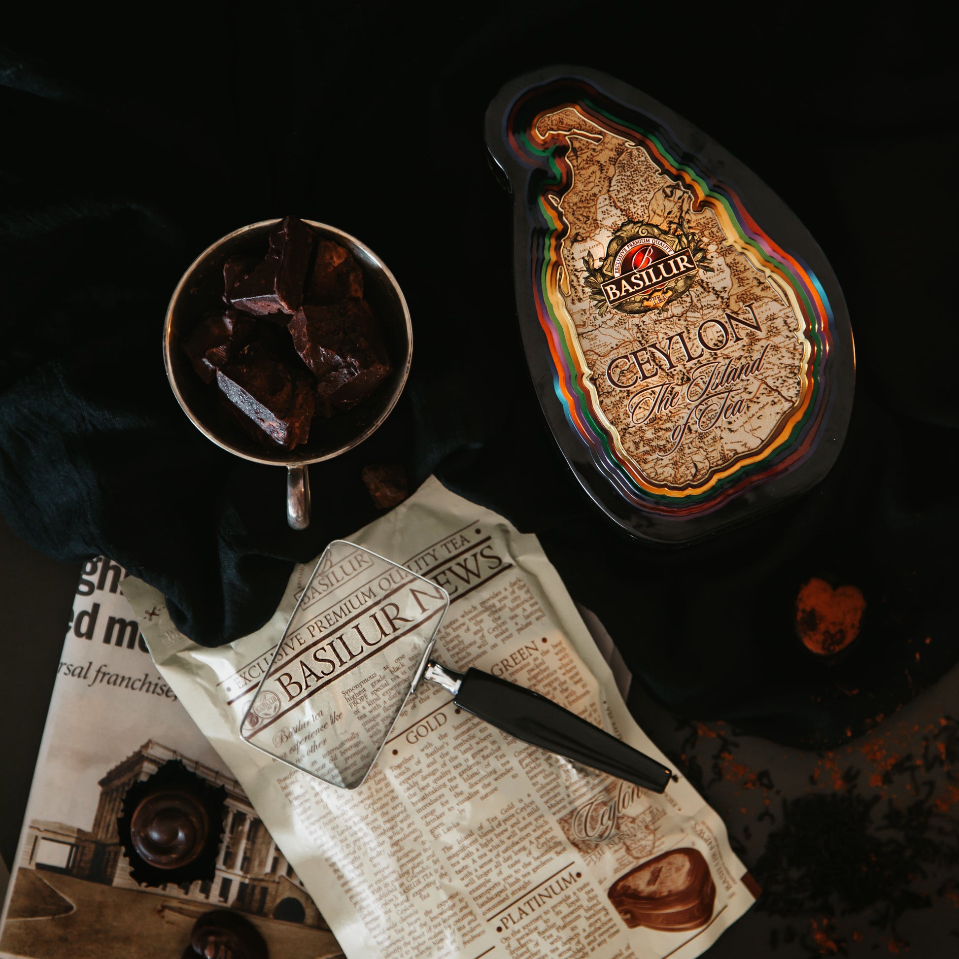 Puszka Basilur Special w otoczeniu czekolady, gazet i akcesoriów na ciemnym tle.