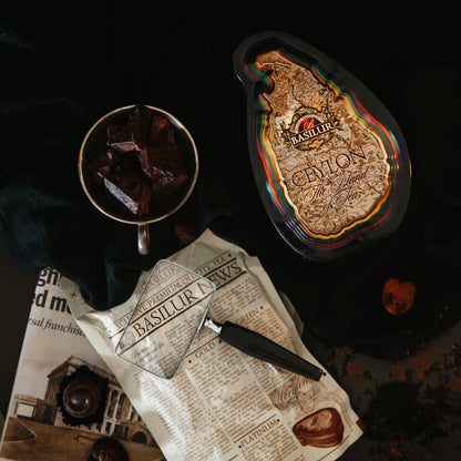 Puszka Basilur Special w otoczeniu czekolady, gazet i akcesoriów na ciemnym tle.