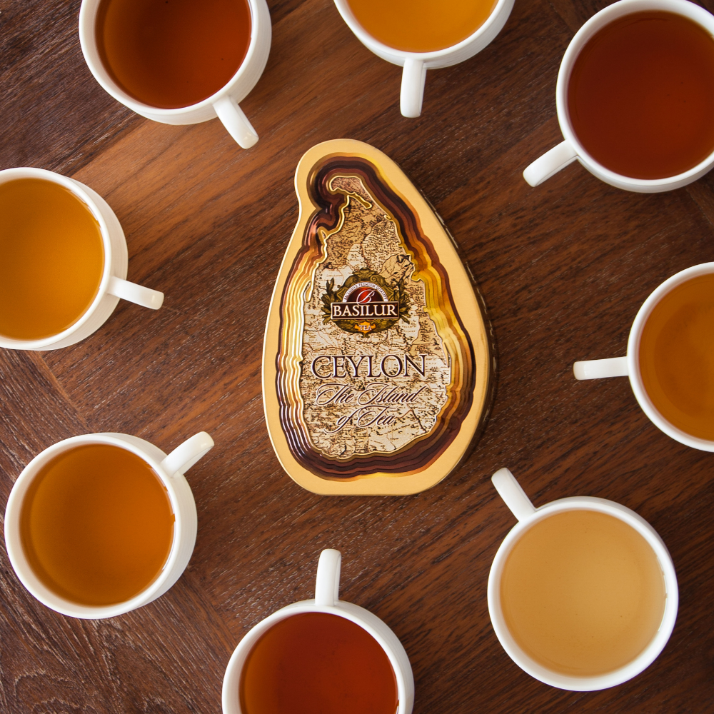 Złota puszka herbaty Basilur Gold otoczona filiżankami z różnymi odcieniami zaparzonej herbaty na drewnianym tle.