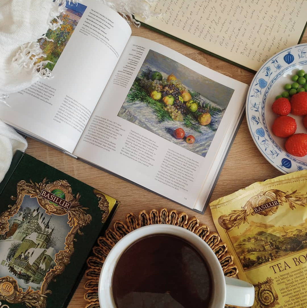 Puszka Basilur Tea Book Vol III obok otwartej książki, filiżanki herbaty i talerzyka z owocami na drewnianym stole.