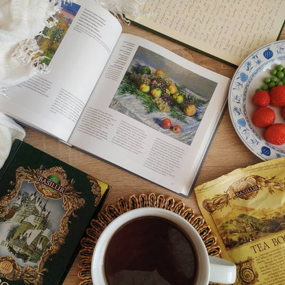 Puszka Basilur Tea Book Vol III obok otwartej książki, filiżanki herbaty i talerzyka z owocami na drewnianym stole.