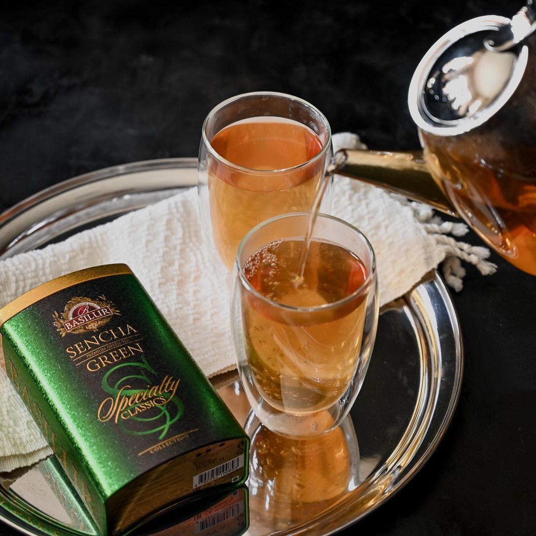 Zielona puszka herbaty Basilur Sencha na tacy obok dwóch szklanek, do których wlewany jest gorący napar.