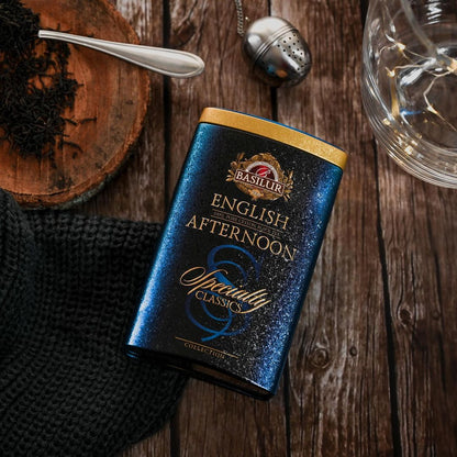 Puszka herbaty Basilur English Afternoon na drewnianym tle obok zaparzacza, filiżanki i liści herbaty.