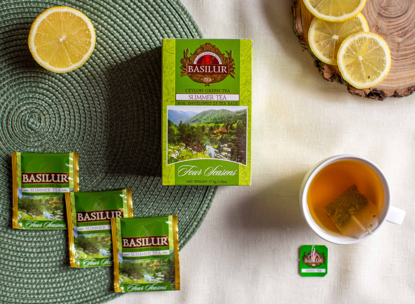 Zestaw herbaty Basilur Summer Tea prezentowany na płaskiej kompozycji. W centrum znajduje się zielone pudełko herbaty Ceylon Green Tea Summer Tea Four Seasons. Obok leżą trzy saszetki w zielonych opakowaniach, filiżanka zaparzonej herbaty z torebką w środku, połówka cytryny oraz plasterki cytryny na drewnianej podstawce. Zdjęcie ma charakter lifestyle i podkreśla letni, świeży aromat herbaty Basilur.
