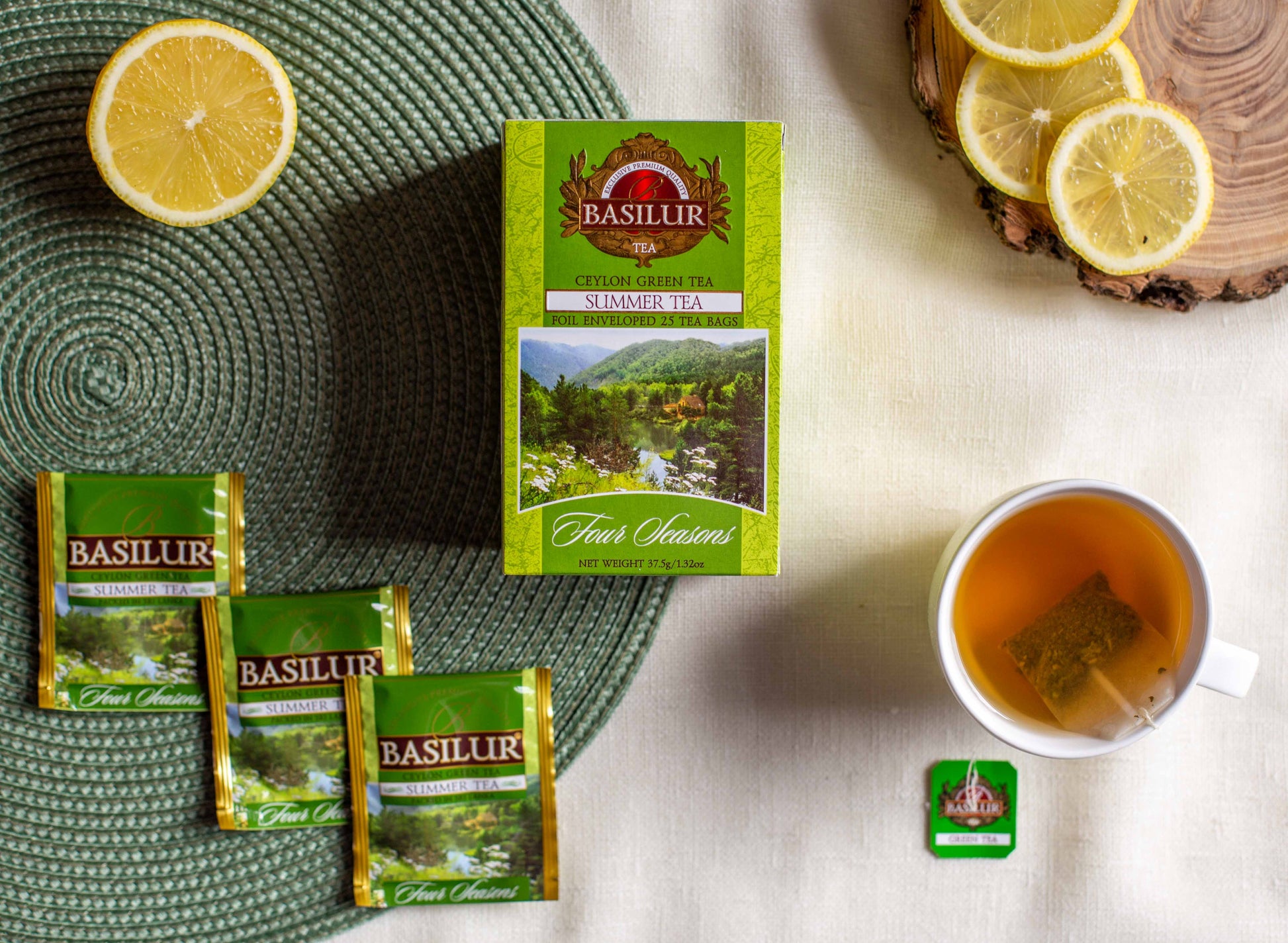 Zestaw herbaty Basilur Summer Tea prezentowany na płaskiej kompozycji. W centrum znajduje się zielone pudełko herbaty Ceylon Green Tea Summer Tea Four Seasons. Obok leżą trzy saszetki w zielonych opakowaniach, filiżanka zaparzonej herbaty z torebką w środku, połówka cytryny oraz plasterki cytryny na drewnianej podstawce. Zdjęcie ma charakter lifestyle i podkreśla letni, świeży aromat herbaty Basilur.