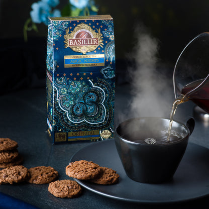Opakowanie herbaty Basilur Magic Nights obok parującej filiżanki czarnej herbaty i ciasteczek.