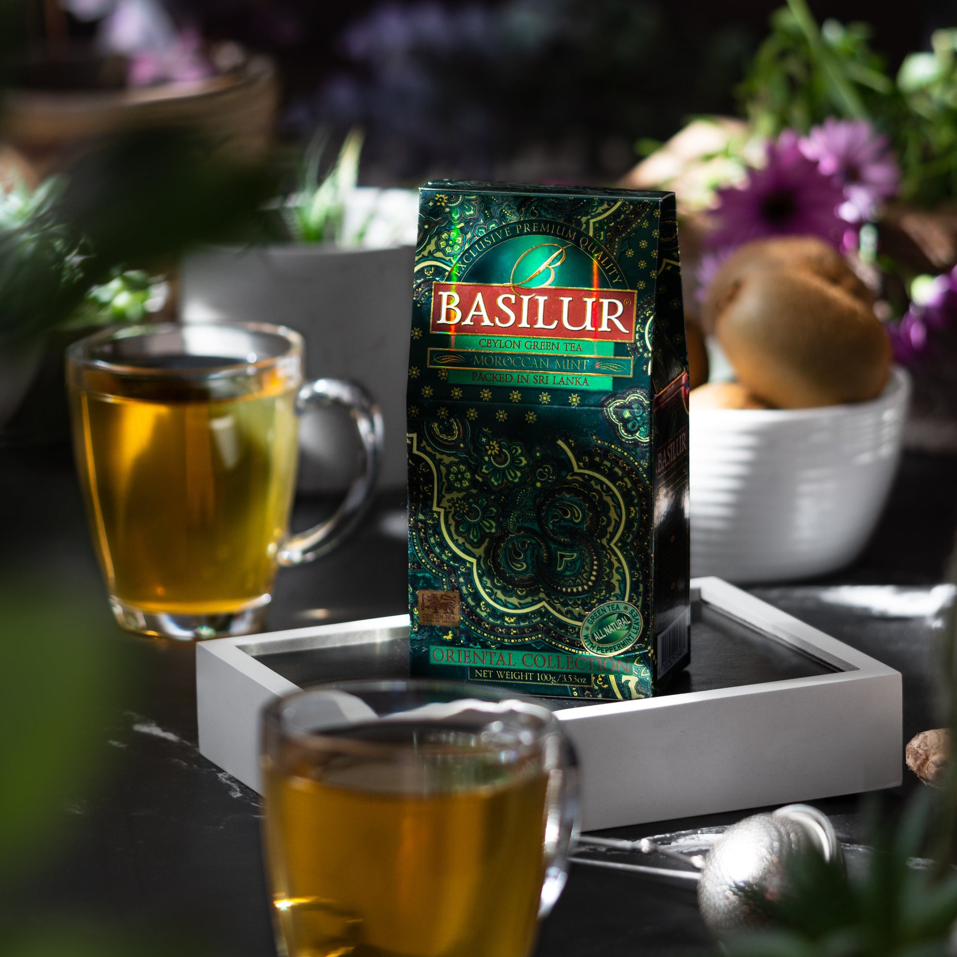 Opakowanie herbaty Basilur Moroccan Mint na tacy w otoczeniu szklanek z gorącą herbatą i świeżych roślin.