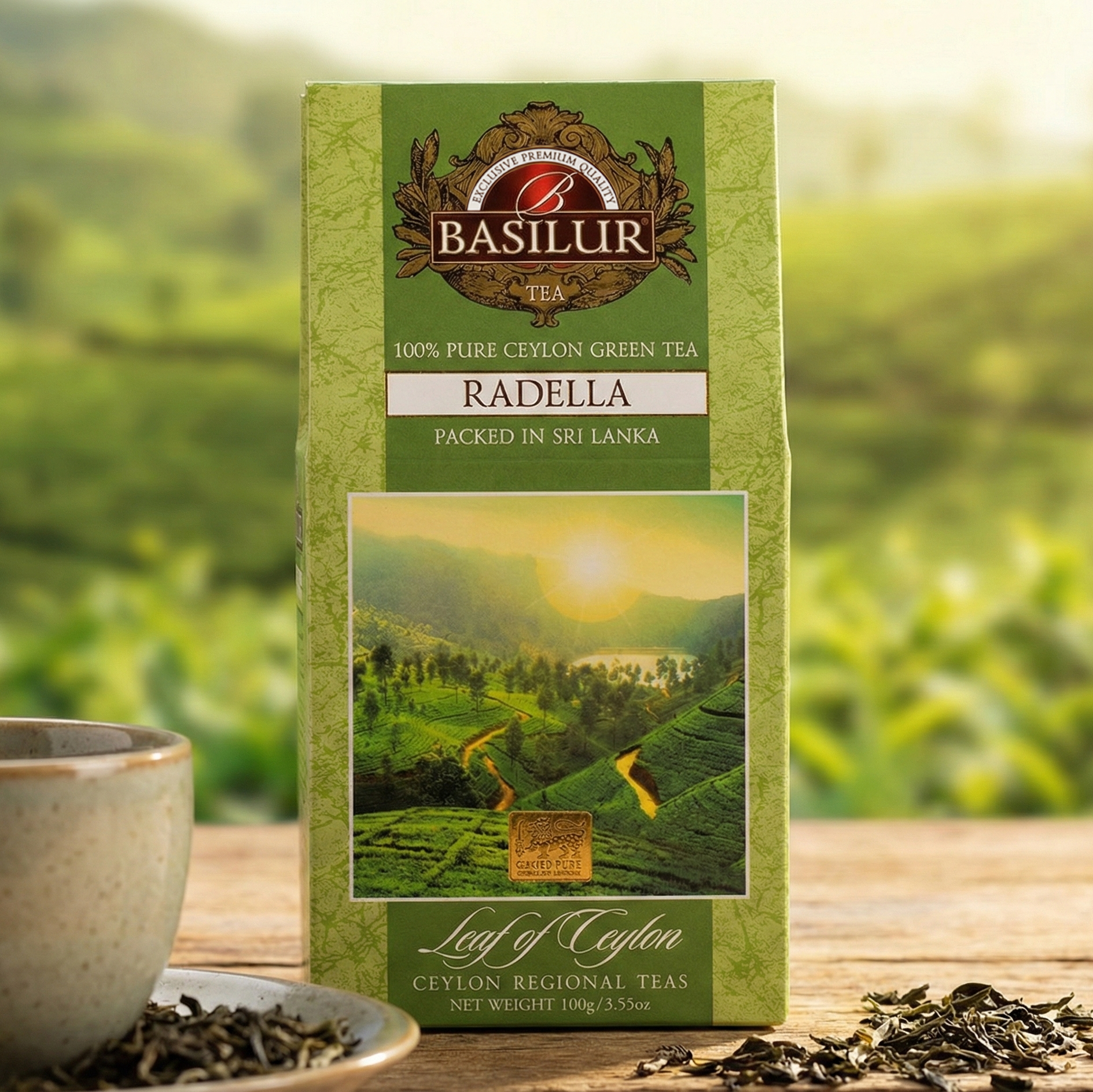 Zielona herbata Basilur Radella 100% Ceylon w opakowaniu, filiżanka i susz na drewnianym stole, w tle plantacja herbaty