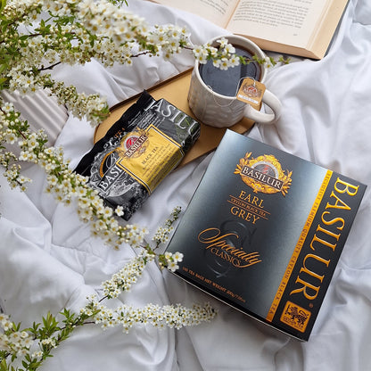 Kubek herbaty Earl Grey Basilur na białej pościeli, obok otwartej książki, opakowania herbaty i kwiatów, spokojny poranek