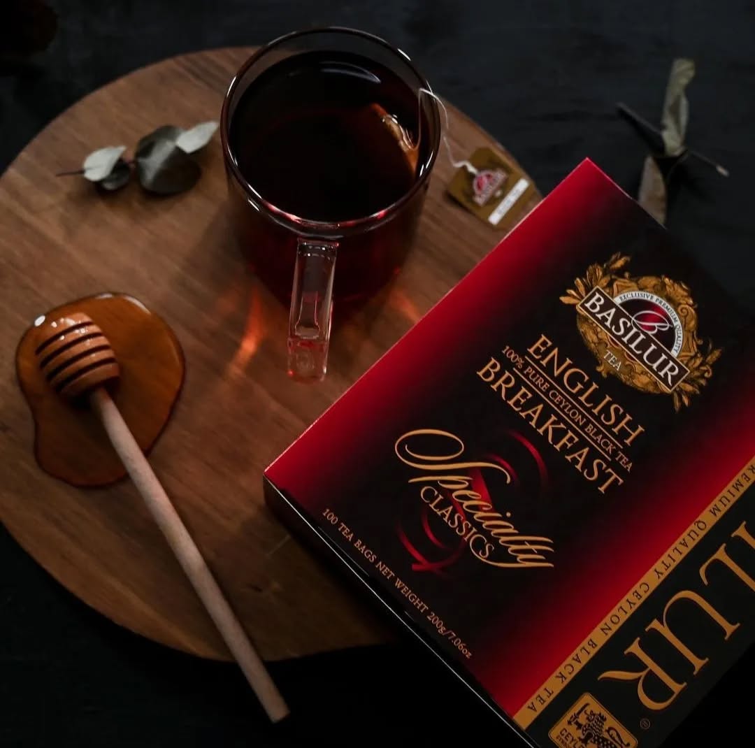 Herbata Basilur English Breakfast w eleganckim opakowaniu, filiżanka czarnej herbaty i drewniany nabierak do miodu na stole