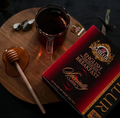 Herbata Basilur English Breakfast w eleganckim opakowaniu, filiżanka czarnej herbaty i drewniany nabierak do miodu na stole