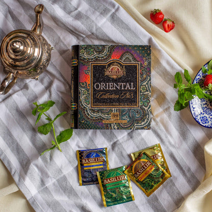 Opakowanie Basilur Oriental Collection No1 ustawione na paskowanym materiale w otoczeniu swiezych truskawek oraz lisci miety. W dolnej czesci widoczne sa kolorowe saszetki roznych wariantow herbat czarnych i zielonych co podkresla orientalny charakter aromatycznych mieszank Basilur.
