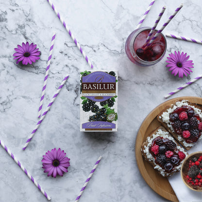 Herbata Basilur Blackcurrant and Blackberry na marmurowym tle obok kanapek z owocami, napoju i fioletowych kwiatów.