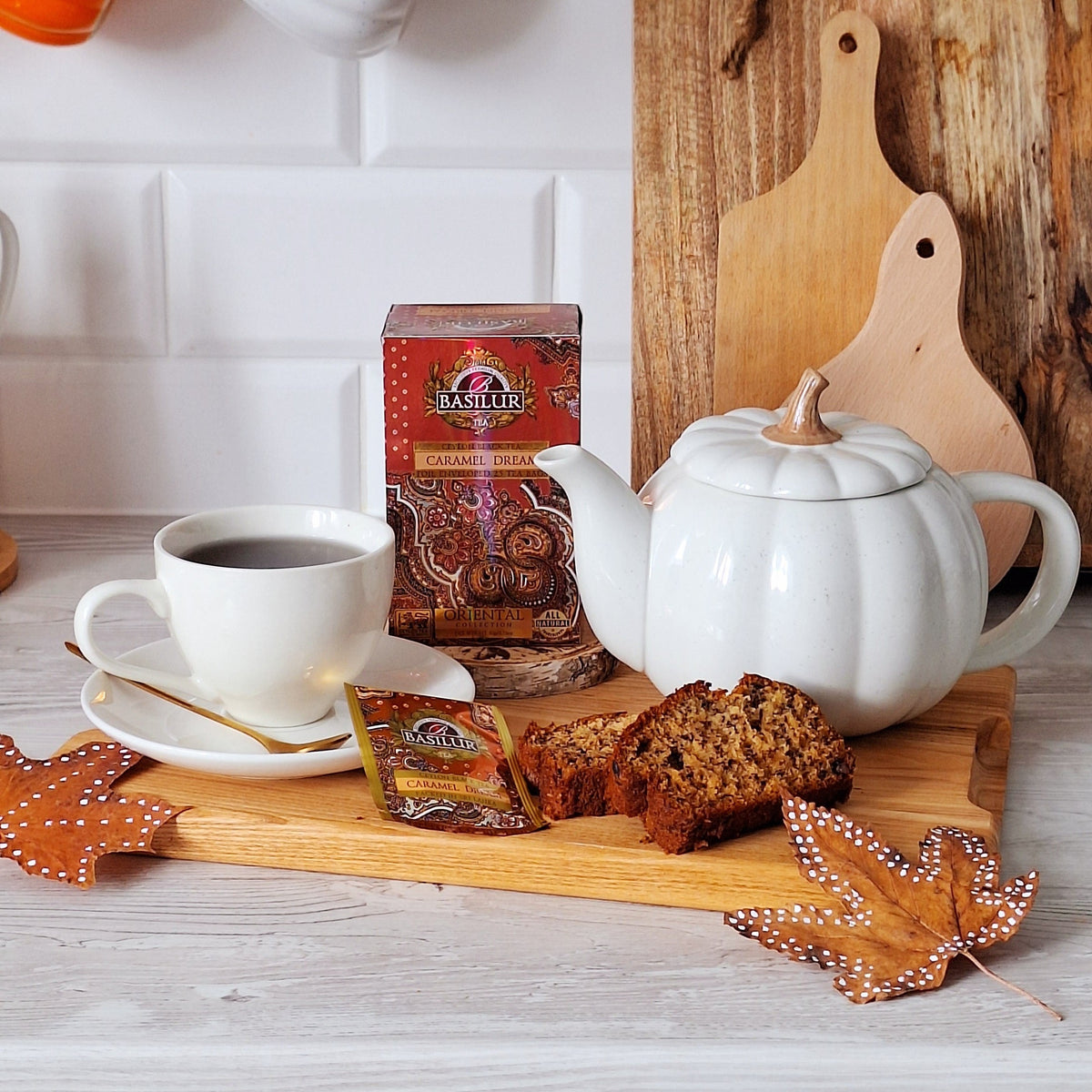 Herbata Basilur Caramel Dream obok filiżanki, białego dzbanka i kawałków ciasta w przytulnej kuchennej aranżacji.