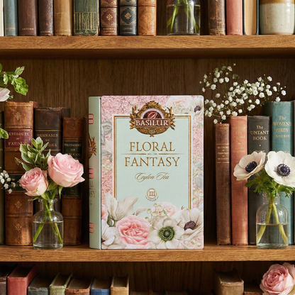 Elegancka herbata Basilur Floral Fantasy w puszce w formie książki, ustawiona na drewnianej półce wśród książek i kwiatów.