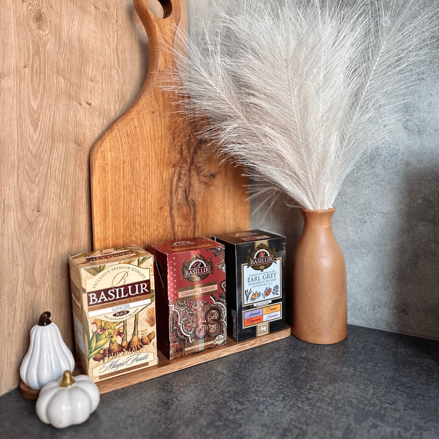Opakowania herbat Basilur Ginger, Caramel Dream i Earl Grey Assorted ustawione przy drewnianych deskach i ozdobnym wazonie.