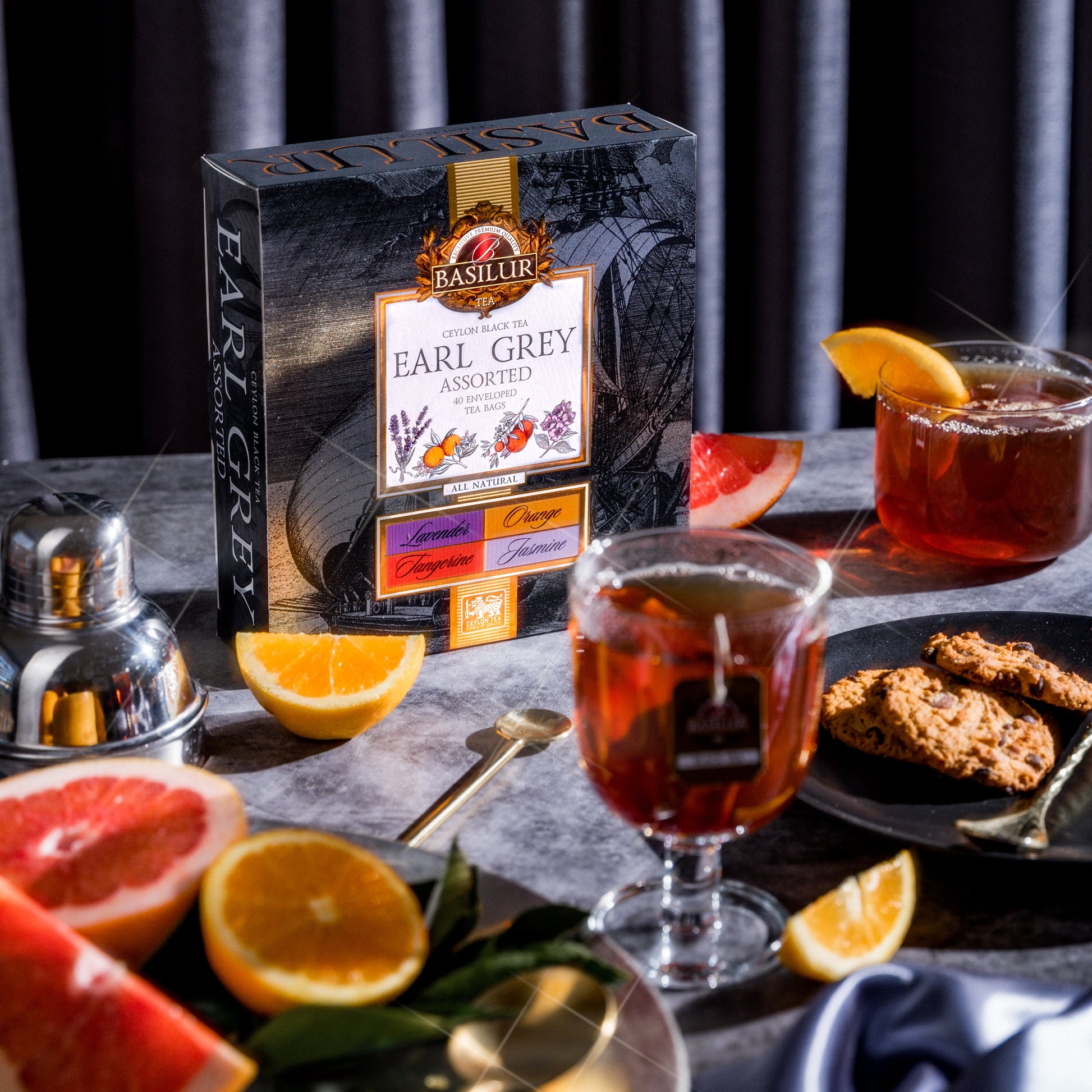 Pudełko herbat Basilur Earl Grey Assorted otoczone cytrusami, herbatą w szklankach i ciasteczkami.