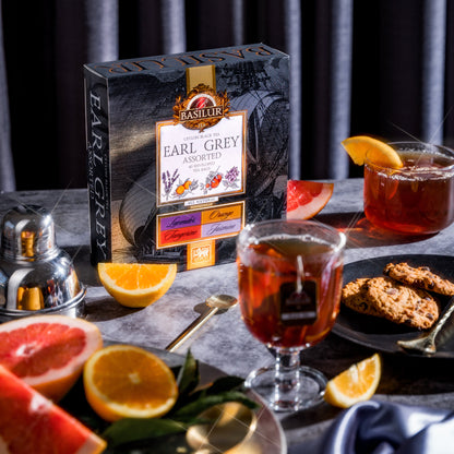 Pudełko herbat Basilur Earl Grey Assorted otoczone cytrusami, herbatą w szklankach i ciasteczkami.
