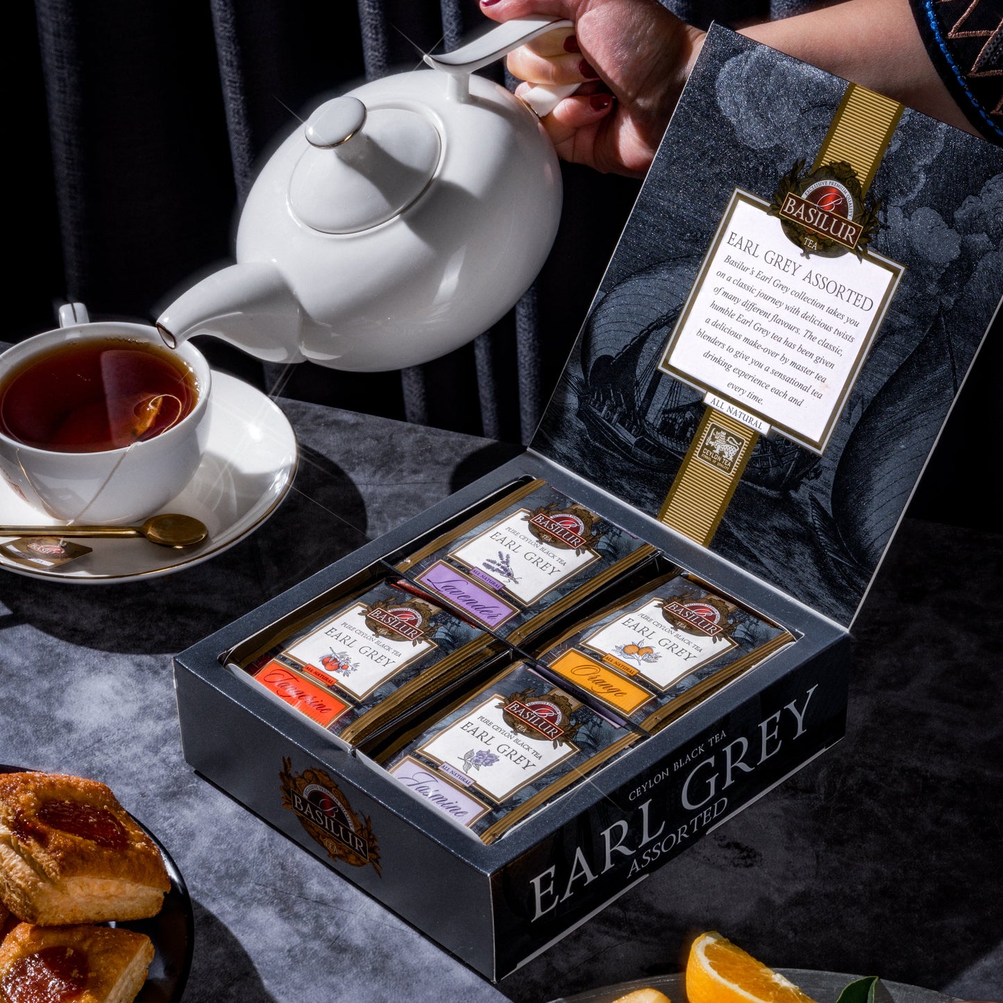 Zestaw herbat Basilur Earl Grey Assorted w otwartym pudełku obok filiżanki z gorącym naparem i owoców cytrusowych.