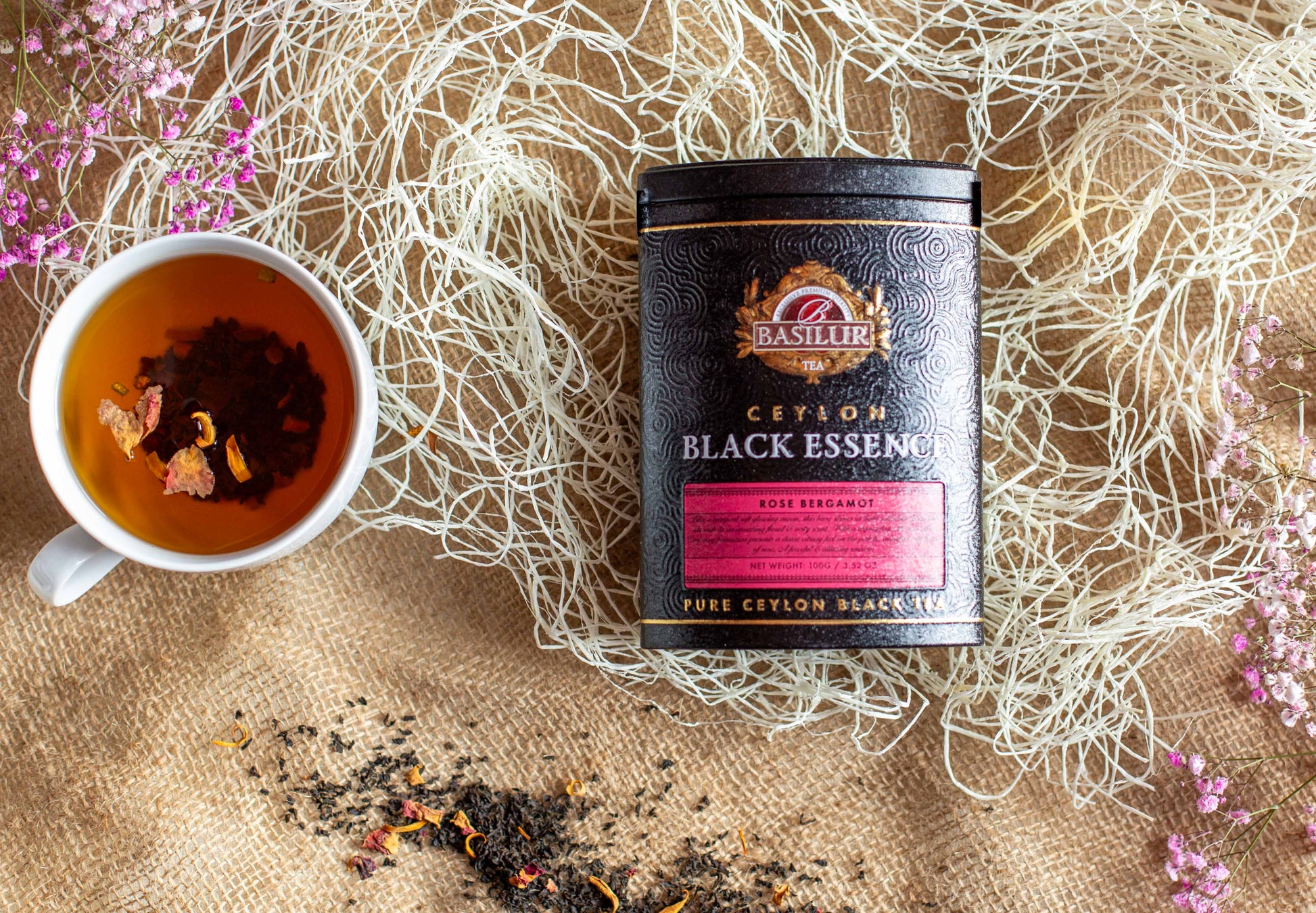 Kompozycja produktowa z czarną herbatą Basilur Black Essence Rose Bergamot w eleganckiej czarno różowej puszce ustawionej na jutowym tle. Obok znajduje się filiżanka zaparzonej czarnej herbaty z widocznymi płatkami róży oraz rozsypane liście herbaty. W tle umieszczono dekoracyjne białe włókna i gałązki różowych kwiatów. Zdjęcie podkreśla aromat róży i bergamotki oraz premium charakter herbaty Basilur.