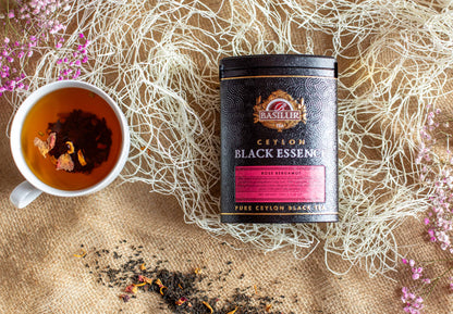 Kompozycja produktowa z czarną herbatą Basilur Black Essence Rose Bergamot w eleganckiej czarno różowej puszce ustawionej na jutowym tle. Obok znajduje się filiżanka zaparzonej czarnej herbaty z widocznymi płatkami róży oraz rozsypane liście herbaty. W tle umieszczono dekoracyjne białe włókna i gałązki różowych kwiatów. Zdjęcie podkreśla aromat róży i bergamotki oraz premium charakter herbaty Basilur.