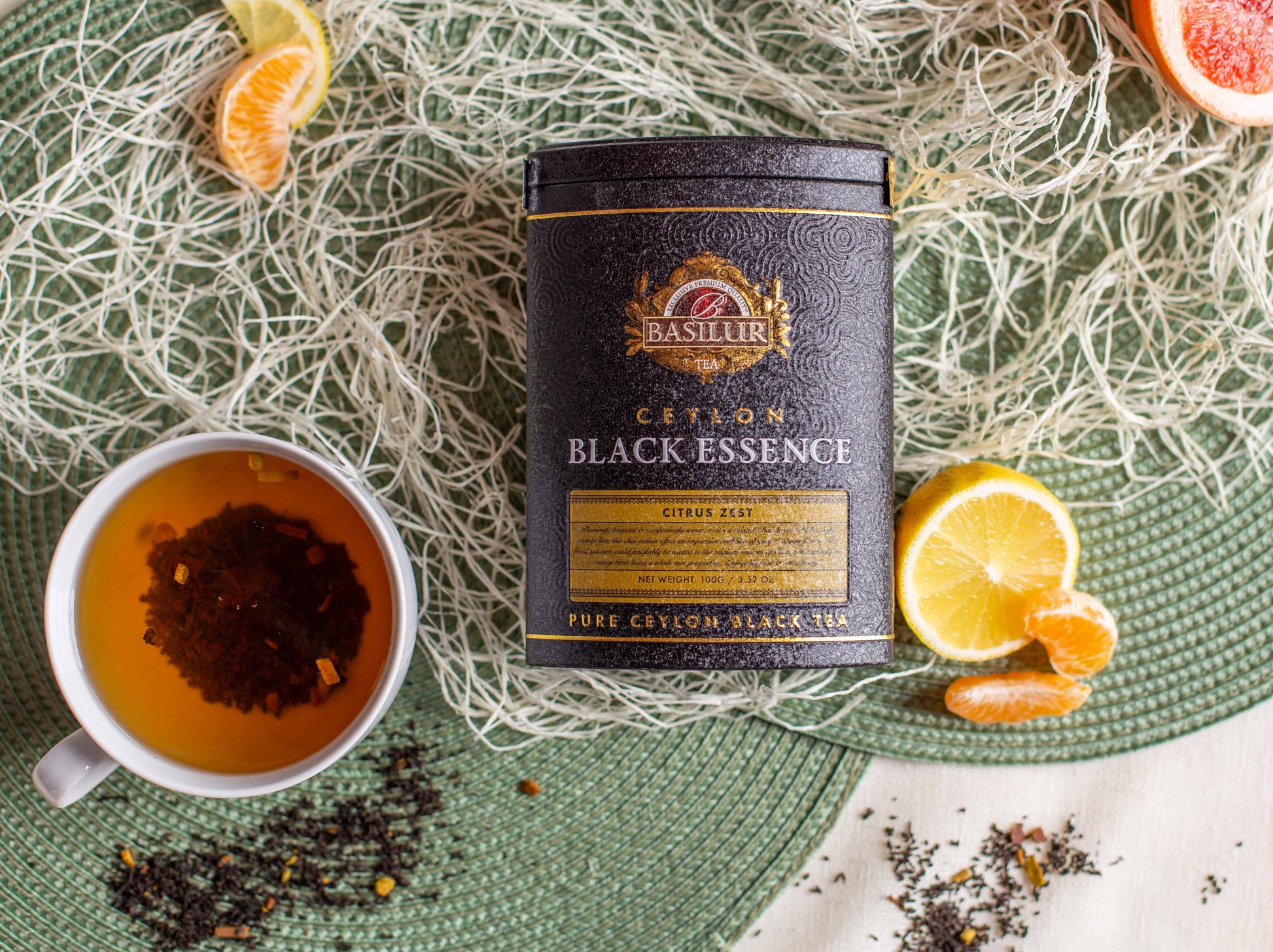 Kompozycja produktowa przedstawiająca czarną herbatę Basilur Black Essence Citrus Zest w eleganckiej czarno złotej puszce ustawionej na zielonej plecionce. Obok znajduje się filiżanka zaparzonej czarnej herbaty z widocznymi cząstkami skórki cytrusowej, rozsypane liście herbaty oraz cząstki mandarynki i połówka cytryny. W tle widoczne są dekoracyjne białe włókna oraz fragment grejpfruta. Zdjęcie podkreśla cytrusowy aromat i luksusowy charakter herbaty Basilur.
