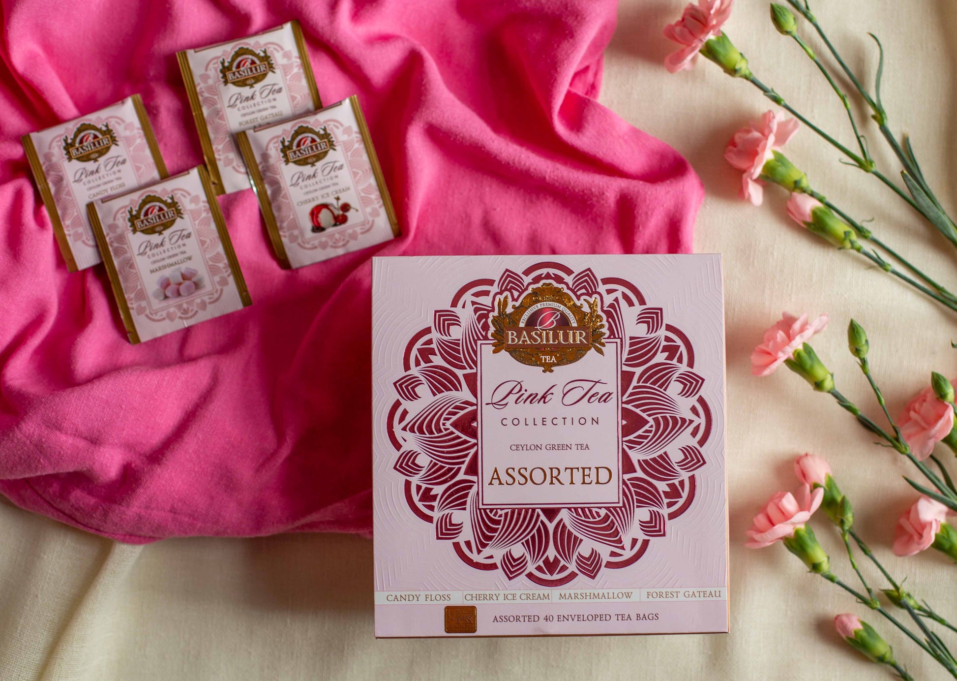 Zamkniete pudelko Basilur Pink Tea Collection Assorted ulozone obok rozowych kwiatow oraz saszetek smakowych na tle intensywnego rozowego materialu. Kompozycja podkresla delikatny dziewczecy i slodki styl kolekcji inspirowanej deserami.