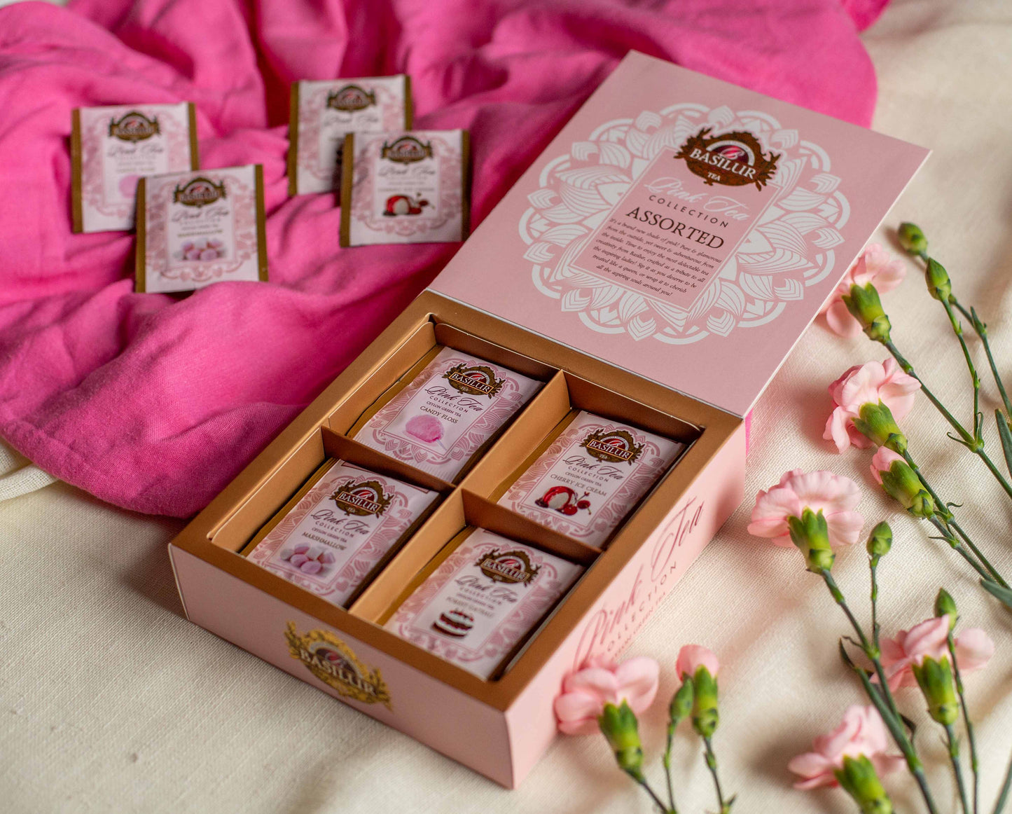 Otwarte opakowanie Basilur Pink Tea Collection Assorted zaprezentowane na rozowym materiale w otoczeniu pastelowych gozdzikow. W srodku widoczne sa saszetki smakowe takie jak candy floss cherry ice cream marshmallow oraz forest gateau co podkresla slodki deserowy charakter zielonej herbaty Ceylon.