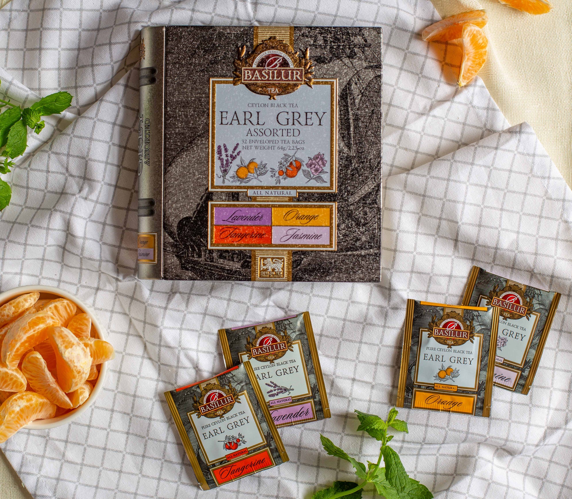 Opakowanie Basilur Earl Grey Assorted ustawione na jasnym materiale w otoczeniu czastek mandarynki oraz lisci miety. Wokół znajduja sie saszetki herbat w smakach takich jak tangerine lavender orange i jasmine co podkresla aromatyczny charakter czarnej herbaty Ceylon i jej naturalne cytrusowo kwiatowe nuty.